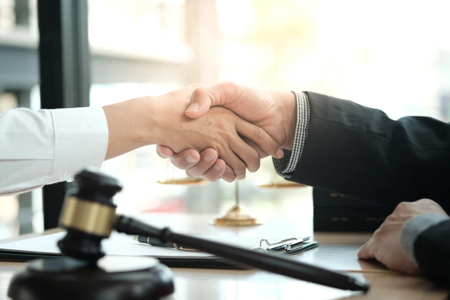 Two people shaking hands over a desk with a gavel, scales of justice, and a clipboard in the foreground.