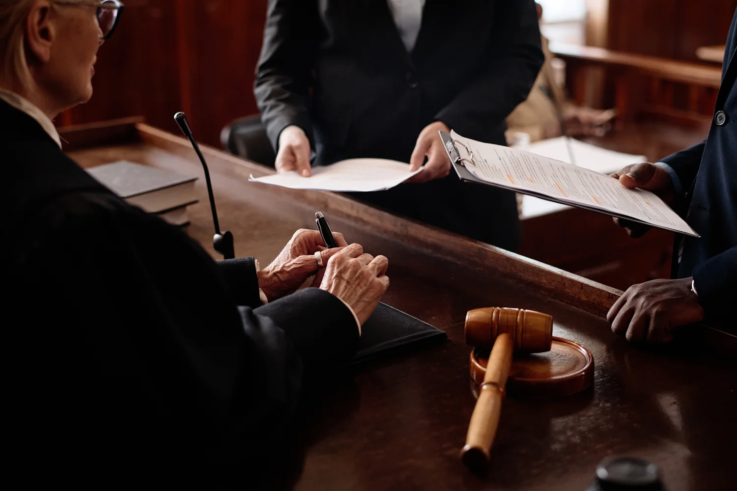 A judge holds a pen while looking at documents handed by a person; a gavel rests on the table.