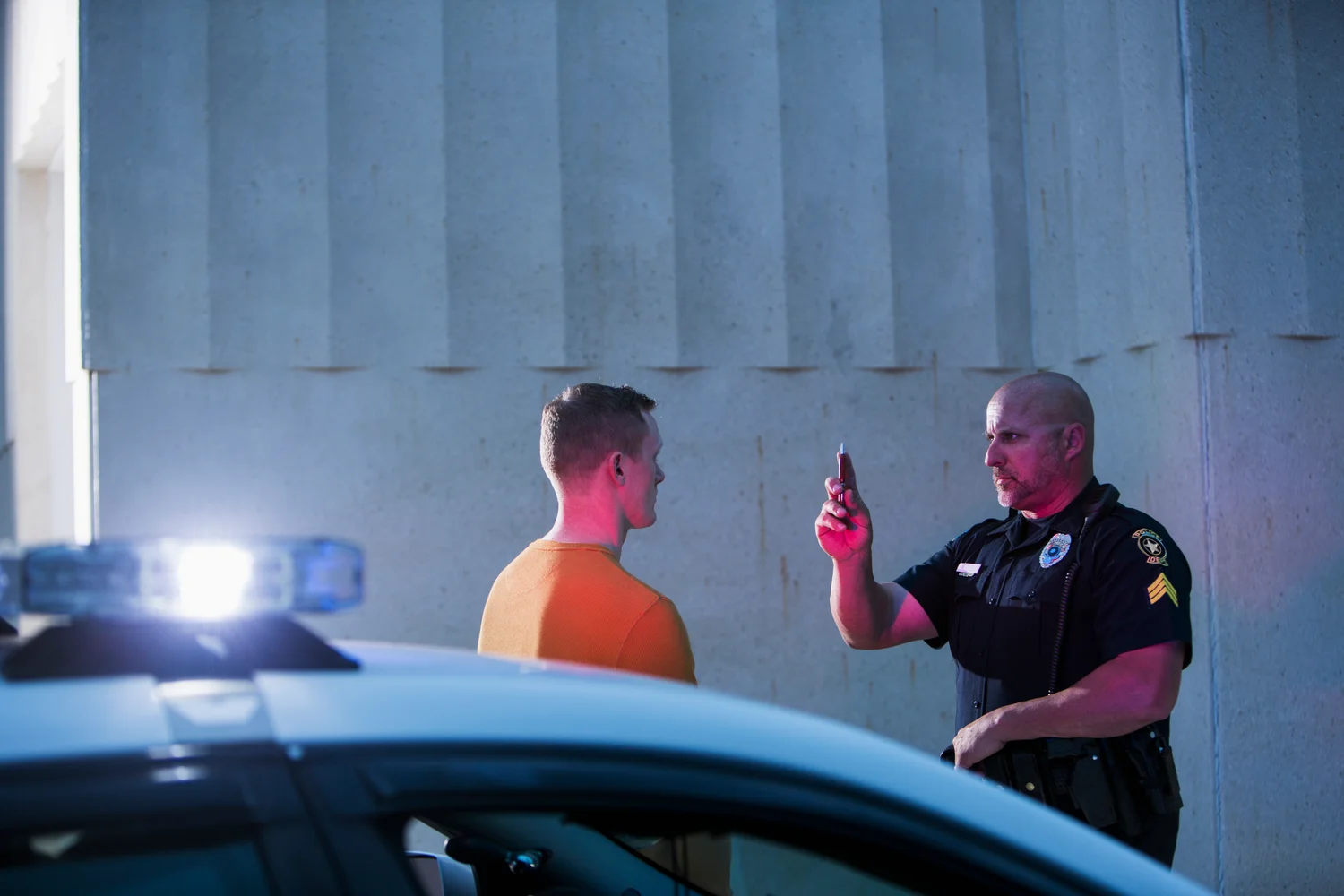 A police officer holds a penlight in front of a man in an orange shirt during a sobriety test. A police car is parked nearby with its lights on.