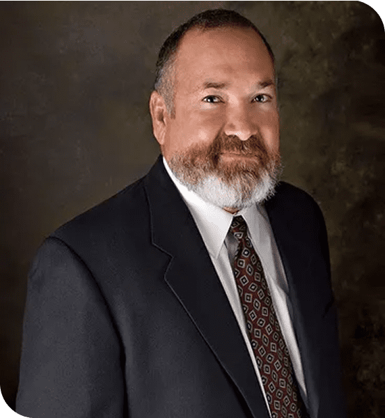 Man with a beard wearing a suit and tie, standing against a dark background, looking at the camera.